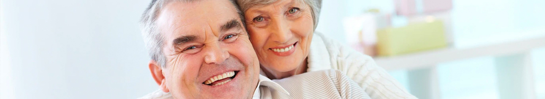 Fotografía en plano detalle de una pareja de la tercera edad abrazados mirando hacia el frente y ambos sonriendo de felicidad.
