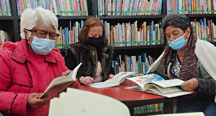 Fotografía de tres mujeres de la tercera edad con tapabocas concentradas leyendo en la biblioteca.