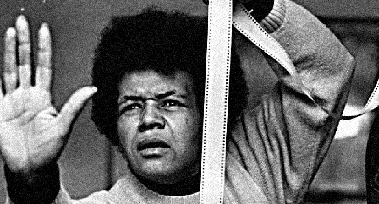 Fotograma de una película de Sarah Maldoror, donde sale ella tomando una película de 35mm, con su mano izquierda en expresión de ¡Para! y su rostros con gesto dramático