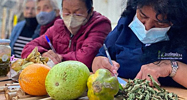 Foto en primer plano de señoras de la tercera edad concentradas tomando apuntes y sobre la mesa frutas y verduras