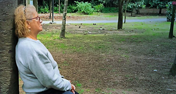 Una señora de la tercera edad con su espalda erguida y sostenida sobre el tronco de un arbol en pose de relajación.