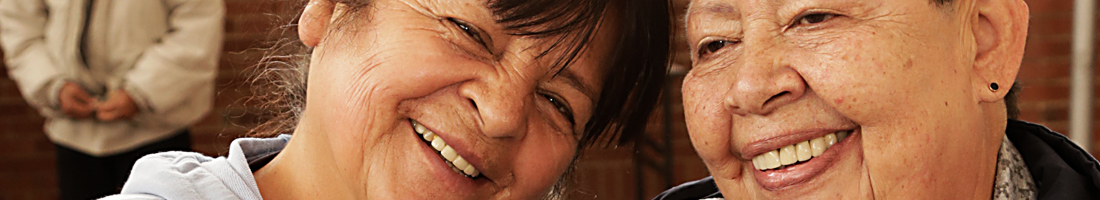 En el primer plano de la fotografía se aprecian dos señoras de la tercera edad felices y sonriendo.