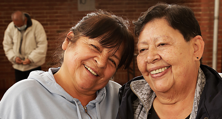 En el primer plano de la fotografía se aprecian dos señoras de la tercera edad felices y sonriendo, al fondo un adulto mayor se encuentra vistiendo una chaqueta