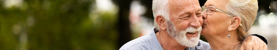 Dos adultos mayores abrazados y la señora le da un beso en la mejilla al señor.