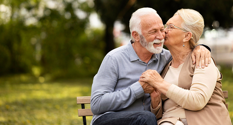Dos adultos mayores abrazados y la señora le da un beso en la mejilla al señor.
