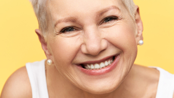 Fotografía primer plano del rostro de una señora de la tercera edad muy sonriente, sobre un fondo de color amarillo.