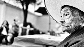 Fotografía en blanco y negro en la cual encontramos en primer plano a una señora de la tercera edad con un sombrero en un espacio exterior que se encuentra desenfocado.