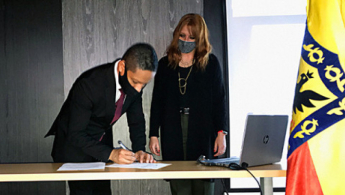 Hombre inclinado hacia adelante, firmando un papel con una mujer a su lado, mirando la acción que realiza.