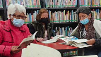 Fotografía de tres mujeres de la tercera edad con tapabocas concentradas leyendo en la biblioteca.