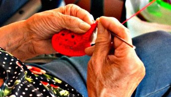 Fotografía en primer plano de las manos de una señora de la tercera edad tejiendo con lana de color rojo.