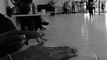 Fotografía en primer plano de unas manos interpretando sobre una conga y de fondo unas bailarinas practicando.