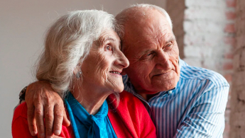 Foto de detalle de dos personas de la tercera edad abrazadas mirando con una sonrisa y de manera esperanzadora hacia una ventana