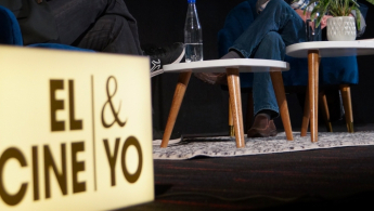 Fotografía en primer plano de una caja de luz donde se proyecta el texto El cine & yo, de fondo se ve en perspectiva un escenario donde se ve claramente que hay unas patas de mesa y se pueden ver una piernas cruzadas.