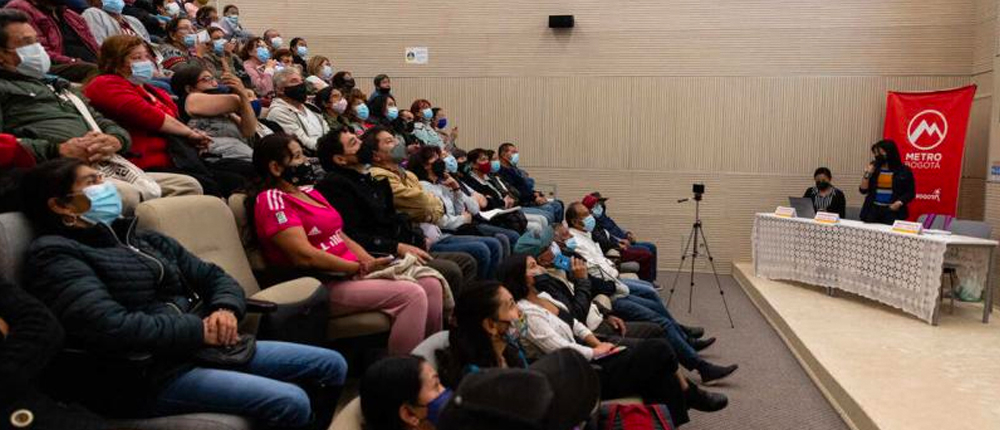 Comunidad reunida en un auditorio en el cual los ponentes se ecuentran hablando y encontramos un pendón con el identificador visual de la marca Metro Bogotá.