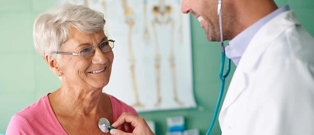 Foto de una señora de la tercera edad siendo atendida por su médico quien le toma signos vitales con el estetoscopio, ambos rien y se encuentran en el consultorio médico.